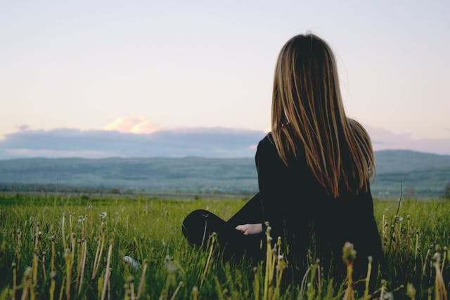 Woman with long hair sitting in the grass looking towards mountains in the distance 