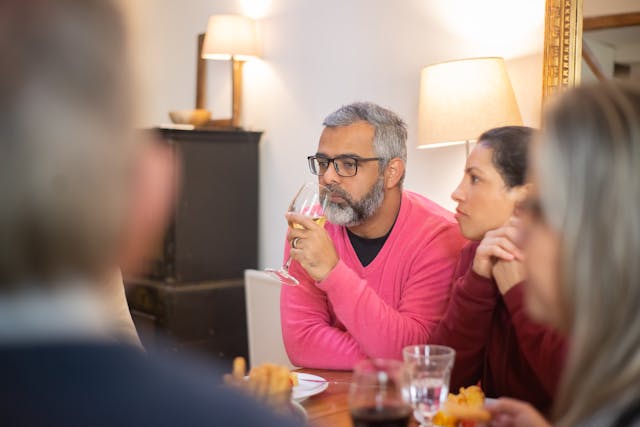 A middle-aged man sitting on a couch among friend while drinking a glass of wine 