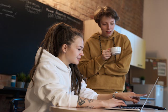 One person sitting in front of a laptop and pointing to something onscreen while another person standing with a cp of coffee looks on 